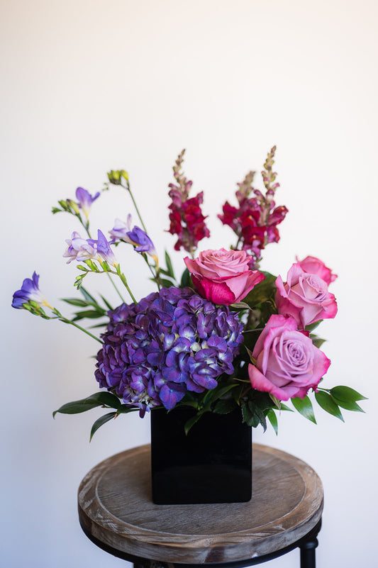 Colorful flower arrangement in a black cube vase on a wooden stool with a white background